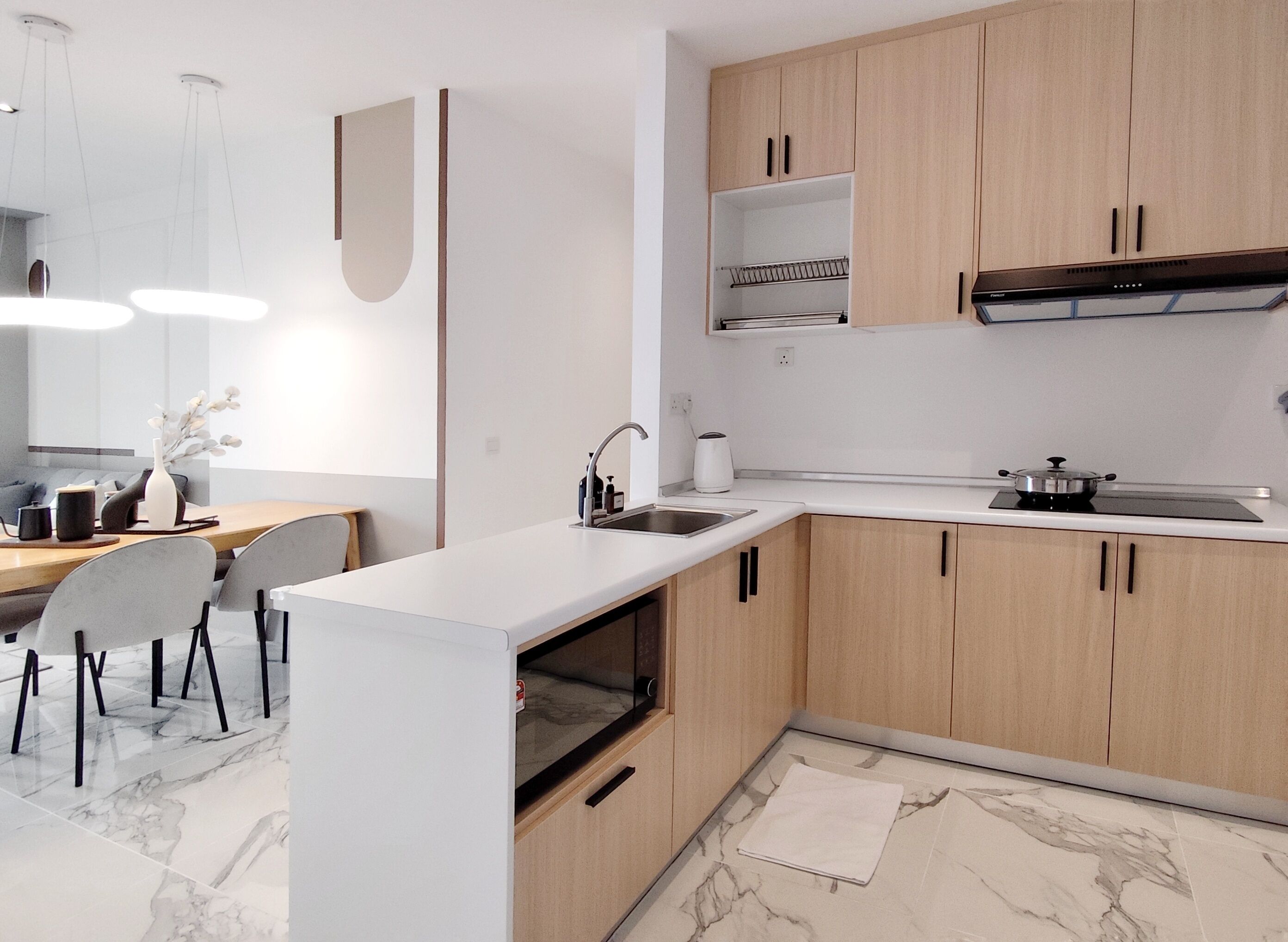 Een moderne keuken met lichte houten kastjes, een wit aanrechtblad en een ingebouwde oven. Er is een eethoek met een marmeren vloer en een witte vaas op tafel.