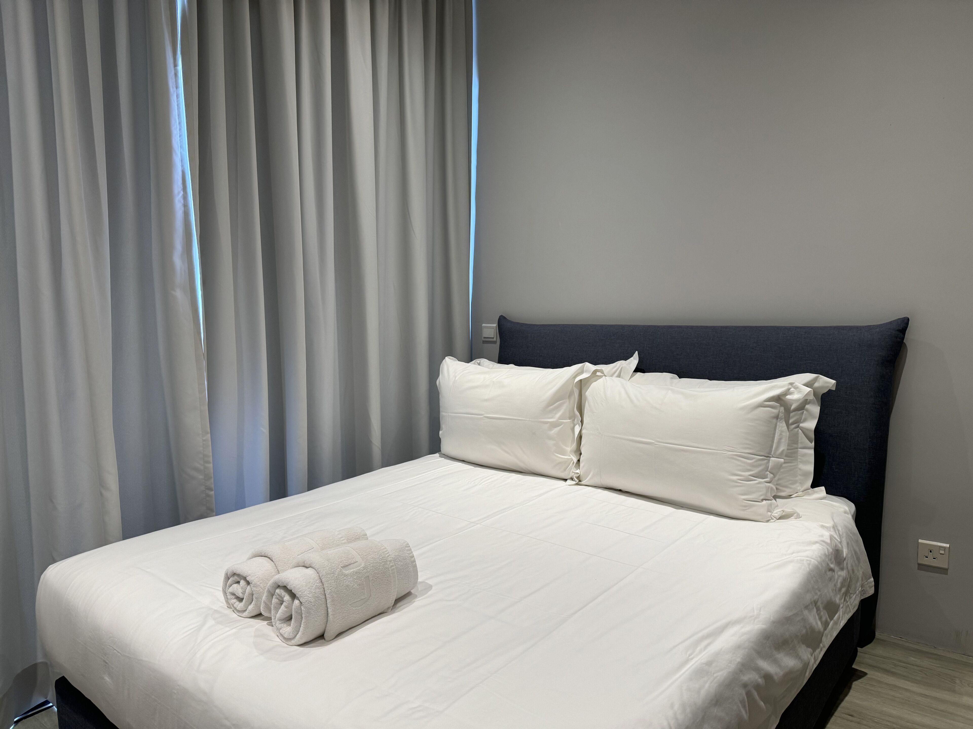 A neatly made bed with white linens and pillows, a dark headboard, and a pair of rolled towels on the foot of the bed.