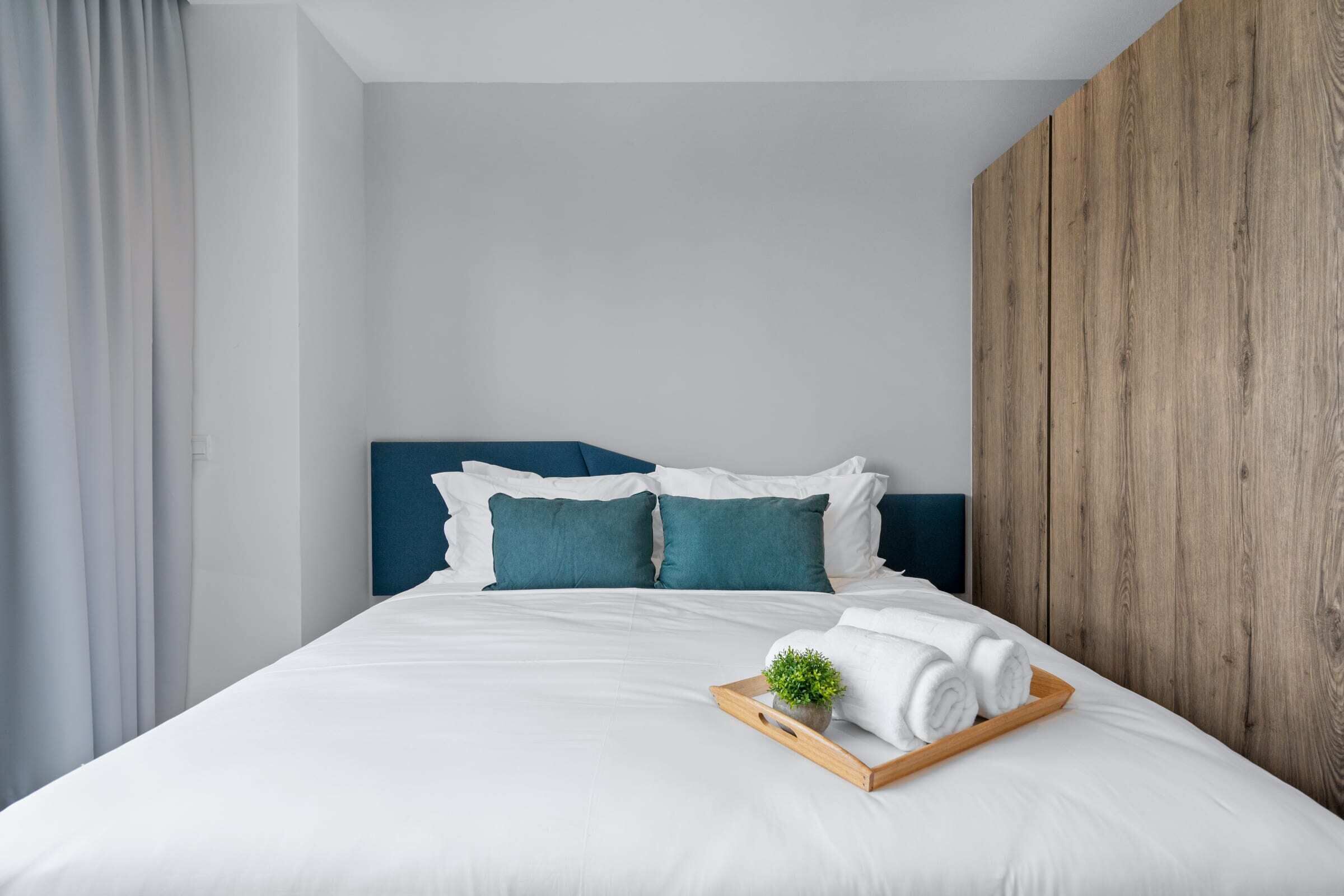 Un lit bien fait, avec une literie blanche et des coussins turquoise, une tête de lit en bois et un plateau contenant des serviettes pliées et une petite plante.