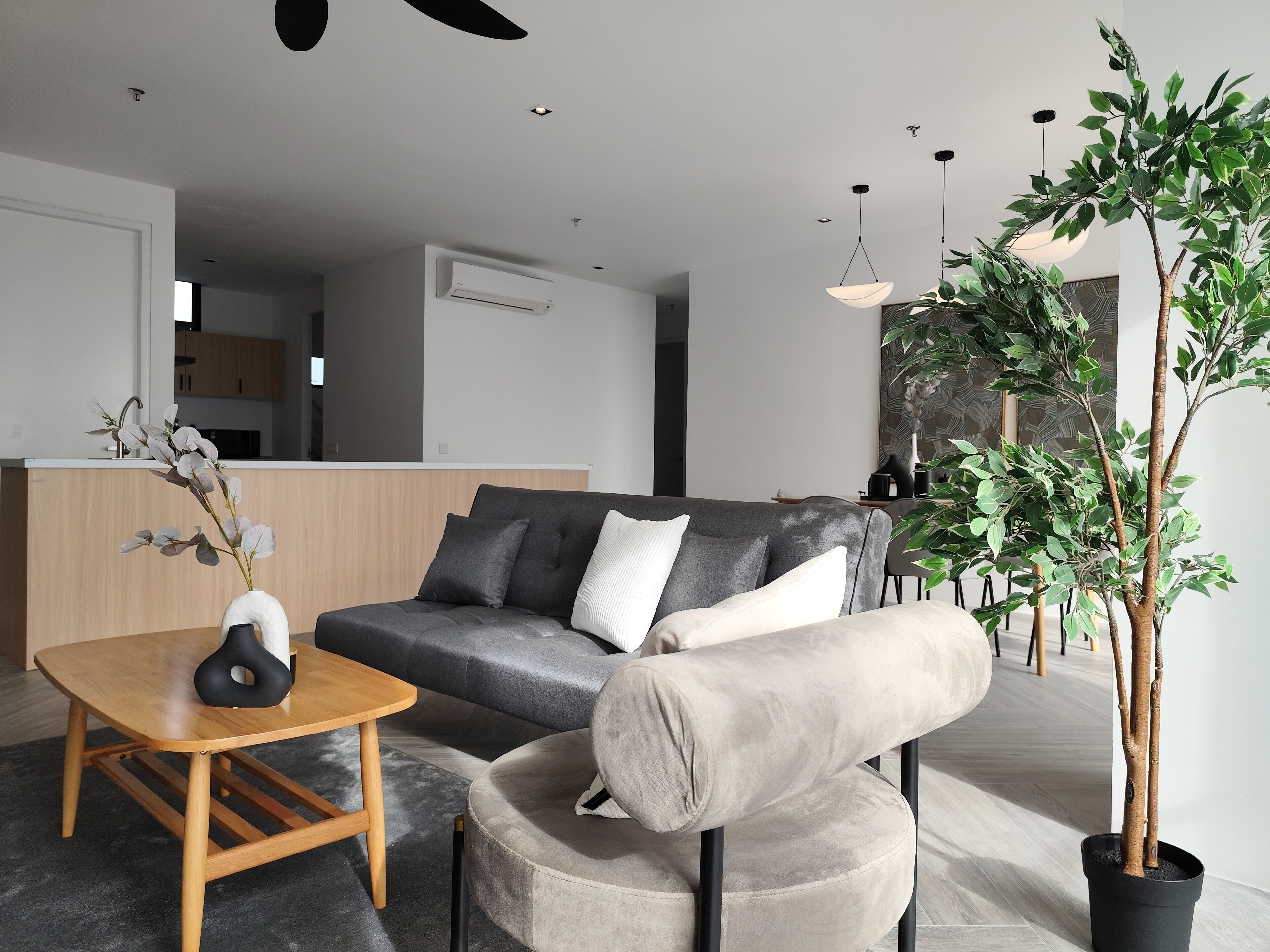 Una sala de estar moderna con un sofá gris, un otoman redondo, una mesa de centro de madera y una planta en maceta.