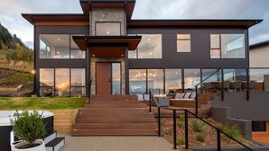 A modern two-story house with a large glass facade, a wooden staircase, and outdoor seating area.
