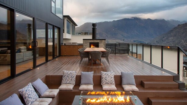 A modern outdoor deck with a fire pit, seating, and a kitchen area overlooking a mountainous landscape.