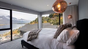 A modern bedroom with a large bed, a view of mountains and a lake, and a hanging copper light fixture.