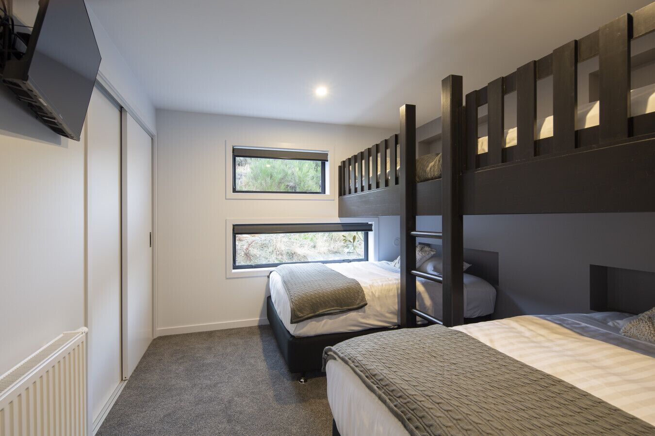A bedroom with bunk beds, two windows, a radiator, and a television mounted on the wall.