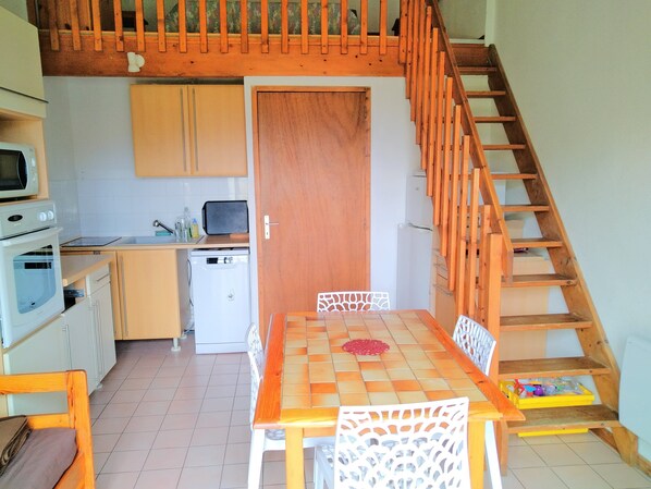 A compact kitchen with a dining area, wooden staircase, and a small refrigerator.