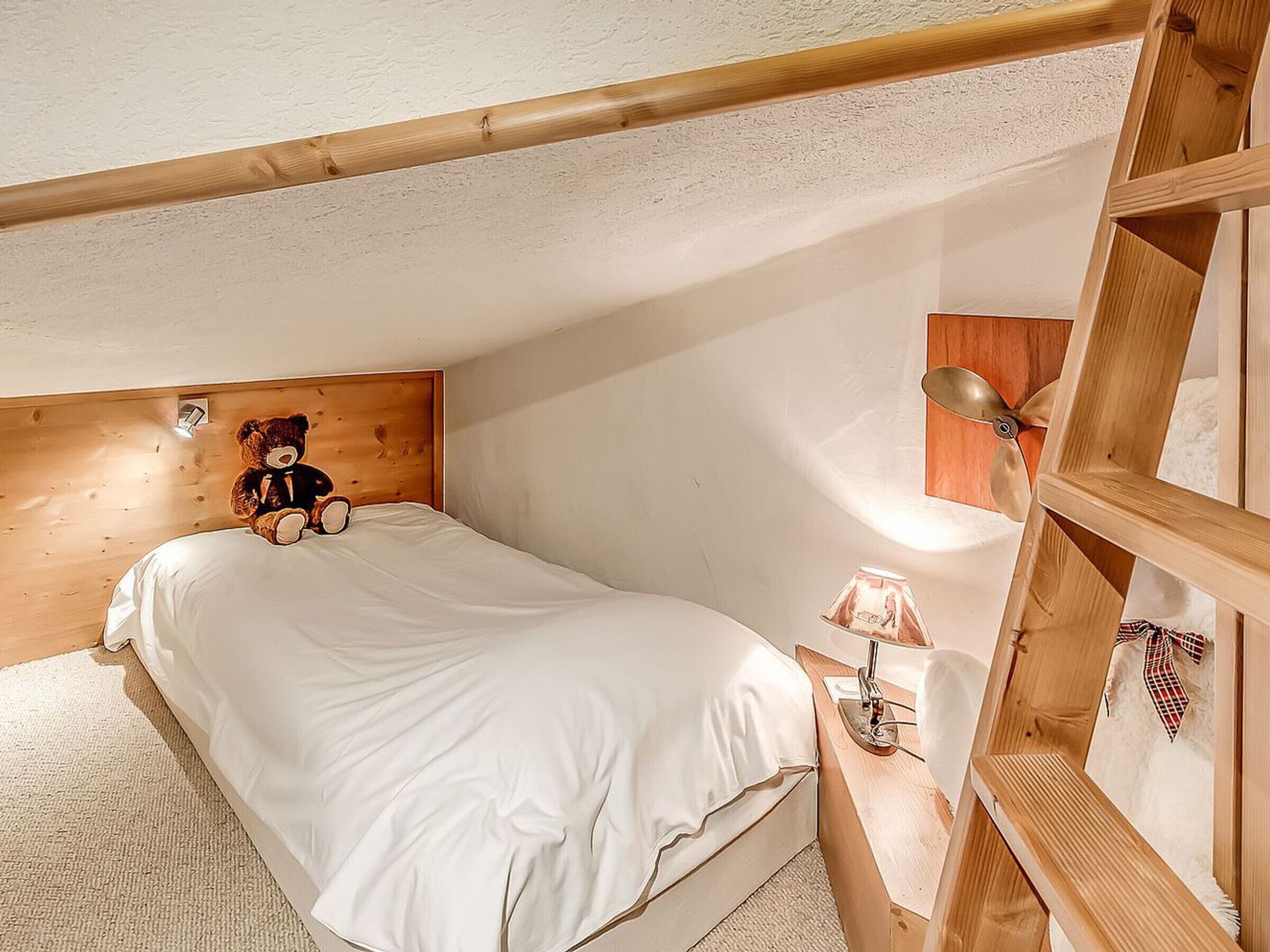 A bedroom with a wooden headboard, a bed with white bedding, a teddy bear, a lamp, and a wooden ladder.