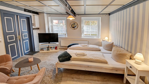 A bedroom with a bed, a TV, a chair, a small table, and a lamp. - fewo1846 - "Unsere Blume" / charming 2 room apartment with balcony / harbor view (Flensburg)