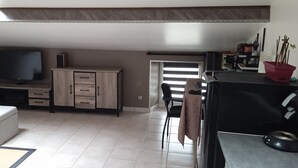 A compact living space with a flat-screen TV, a wooden cabinet with drawers, a sofa, a dining table with chairs, and a small kitchen area with a microwave and a red pot on the counter. - Apartment (NERAC)