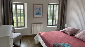 A bedroom with a bed, a chair, two windows, a radiator, and a painting on the wall. - House (Agon-Coutainville)