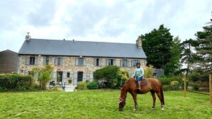 A stone building with a grey roof, a person riding a horse in a grassy field, and a wooden fence in the background. - House (Agon-Coutainville)