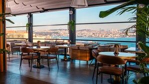 Un restaurant moderne avec des tables et des chaises rondes, de grandes fenêtres donnant sur la ville et des plantes d’intérieur.