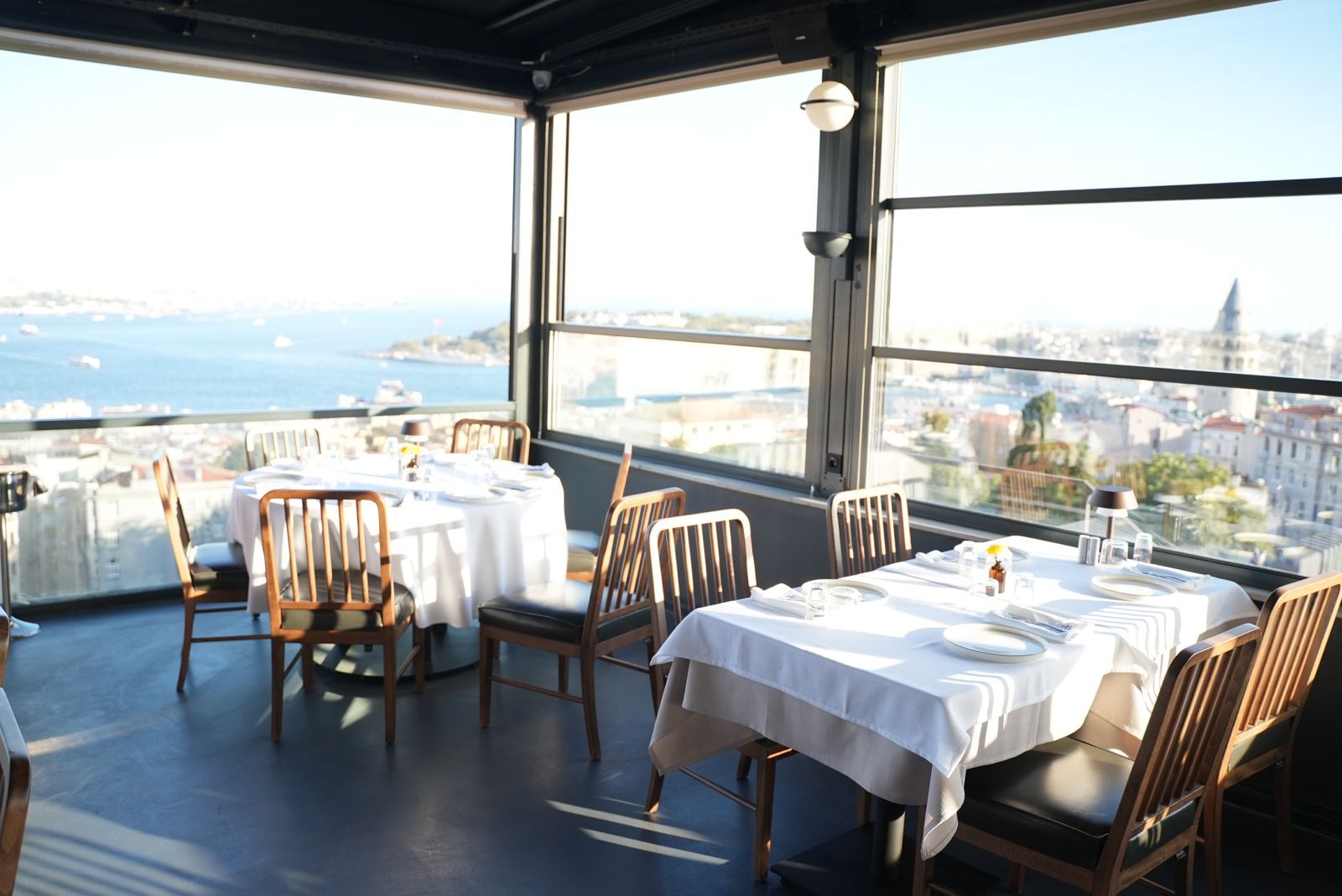 Un restaurante con mesas preparadas para comer, amplios ventanales que ofrecen vistas a la ciudad y al mar, y una decoración de diseño moderno.