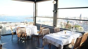 Un restaurante con mesas preparadas para comer, amplios ventanales que ofrecen vistas a la ciudad y al mar, y una decoración de diseño moderno.