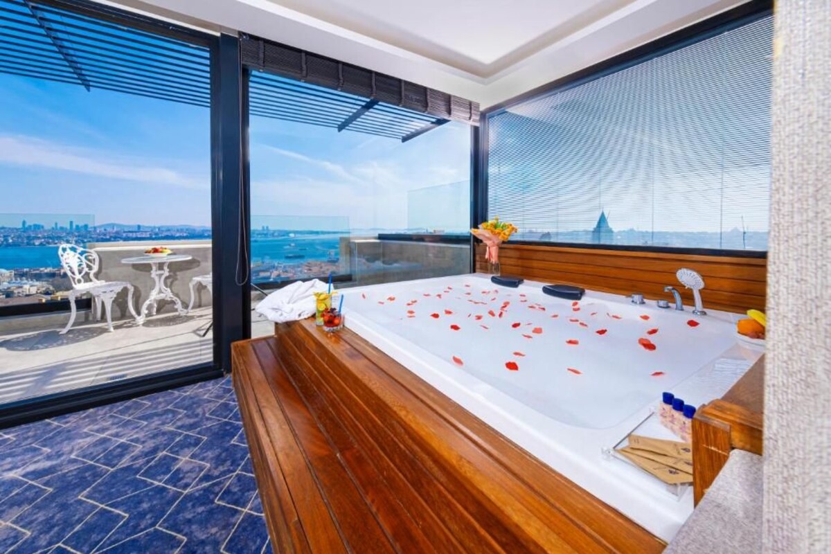 A modern bathroom with a large bathtub, a wooden bench, and a view of the city through large windows.