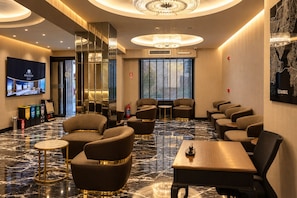 Un hall d’hôtel au design moderne, avec un grand lustre, un espace salon comprenant des canapés et des tables, et un écran de télévision qui affiche une image d’une chambre.
