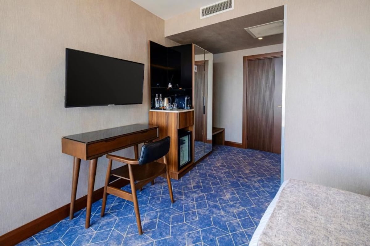 A hotel room with a flat-screen TV mounted on the wall, a wooden desk with a chair, a minibar, and a wardrobe.
