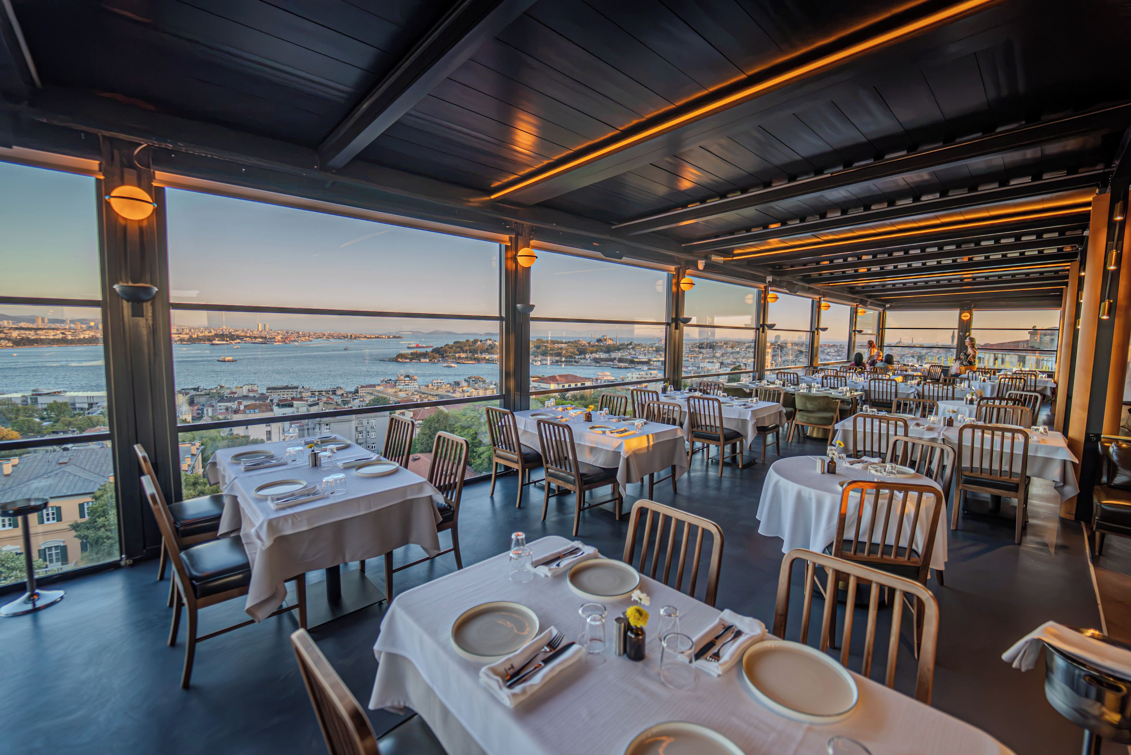 A restaurant with tables set for dining, large windows offering a view of a cityscape and water.