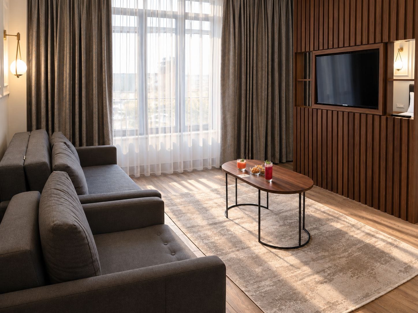 A modern living room with a sofa set, a coffee table with drinks, a flat-screen TV, and a view through large windows.