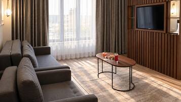 A modern living room with a sofa set, a coffee table with drinks, a flat-screen TV, and a view through large windows.