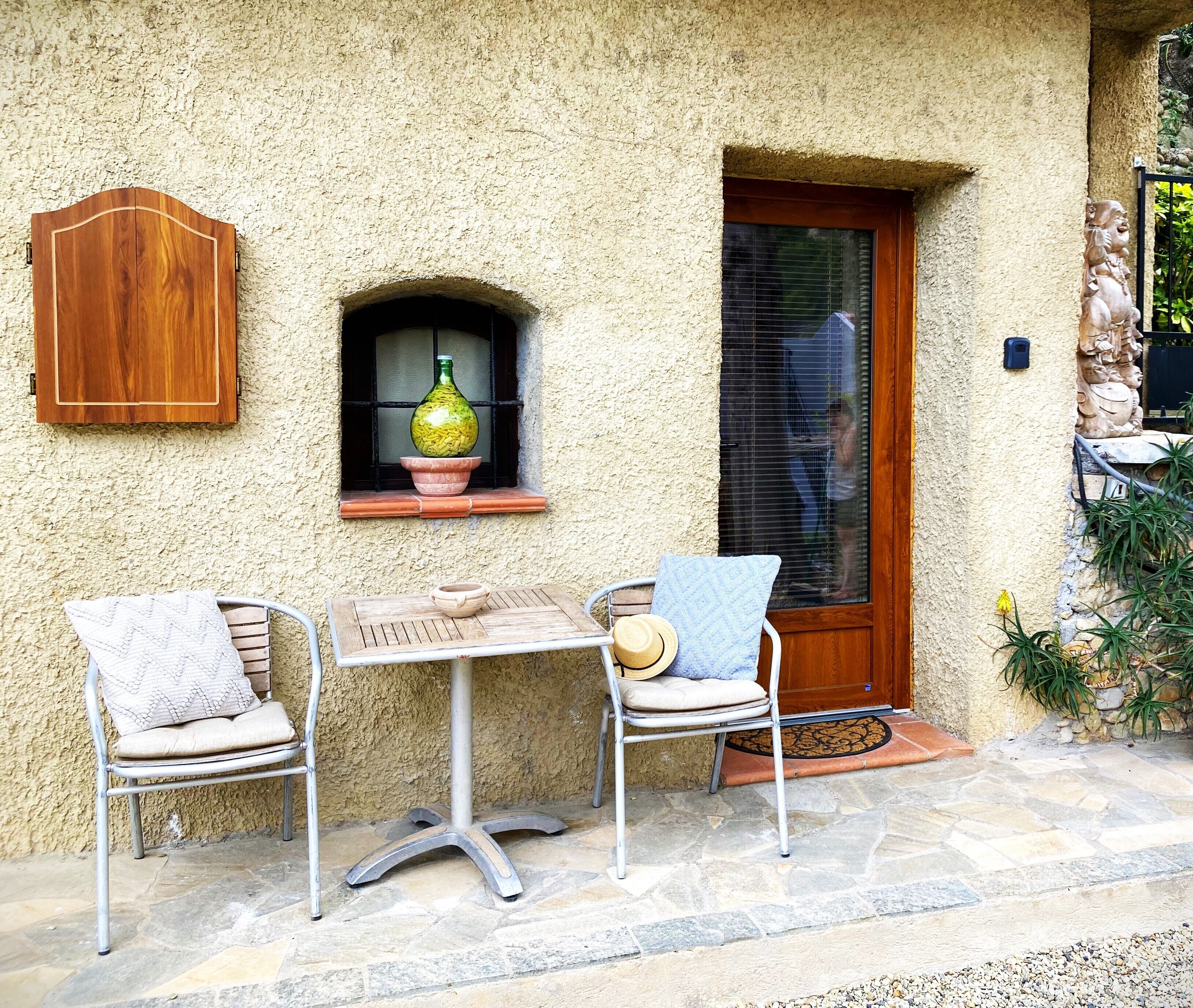 Eine Terrasse mit Tisch und zwei Stühlen, einem Holzkasten, einem Fenster mit Flasche und Schale sowie einer Tür mit Fußmatte.