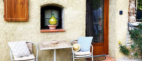 Eine Terrasse mit Tisch und zwei Stühlen, einem Holzkasten, einem Fenster mit Flasche und Schale sowie einer Tür mit Fußmatte.