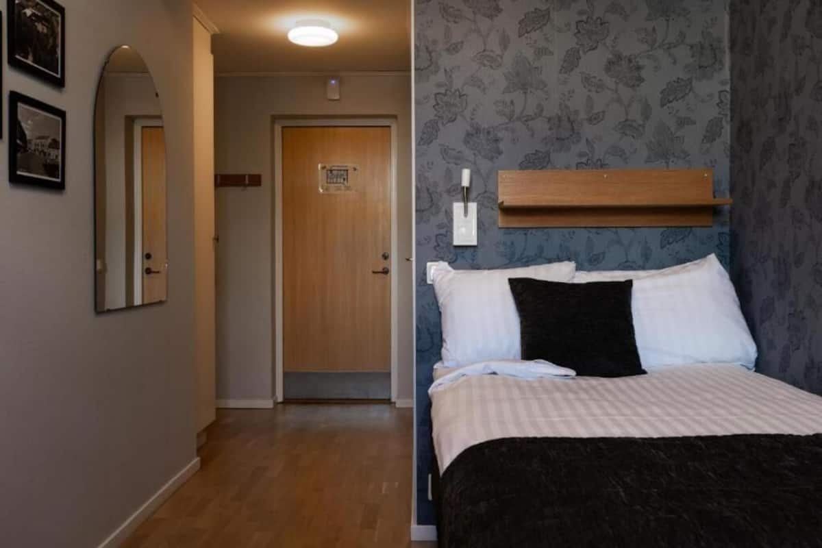 Quarto de hotel com cama, porta de madeira, interruptor de luz e prateleira fixada na parede.