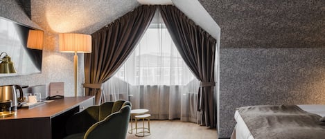 A modern hotel room with a desk, two chairs, a bed, and a large window with curtains.