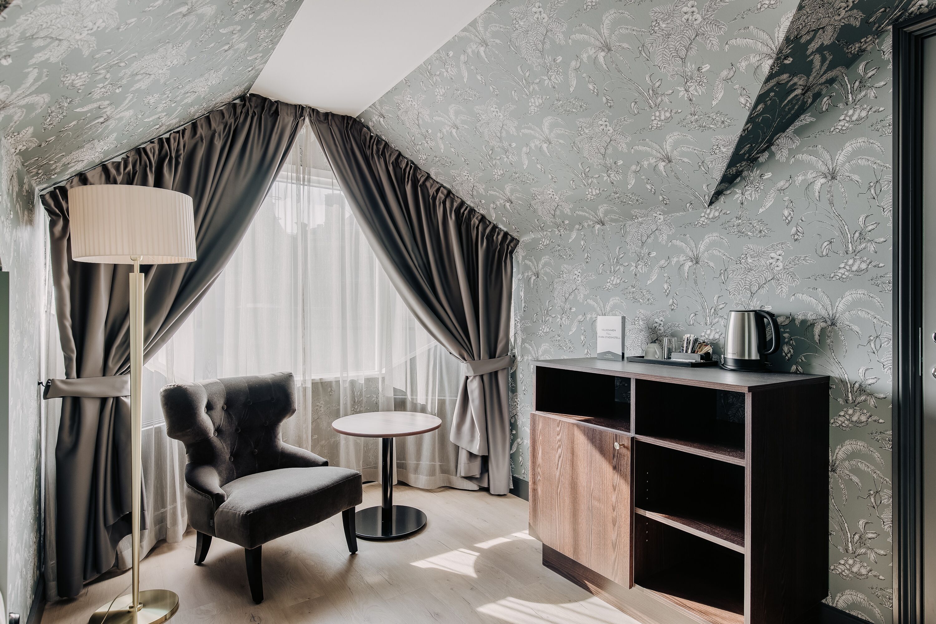 Una habitación con un sillón oscuro, una mesita redonda pequeña, una lámpara de pie y un aparador de madera con estantes.