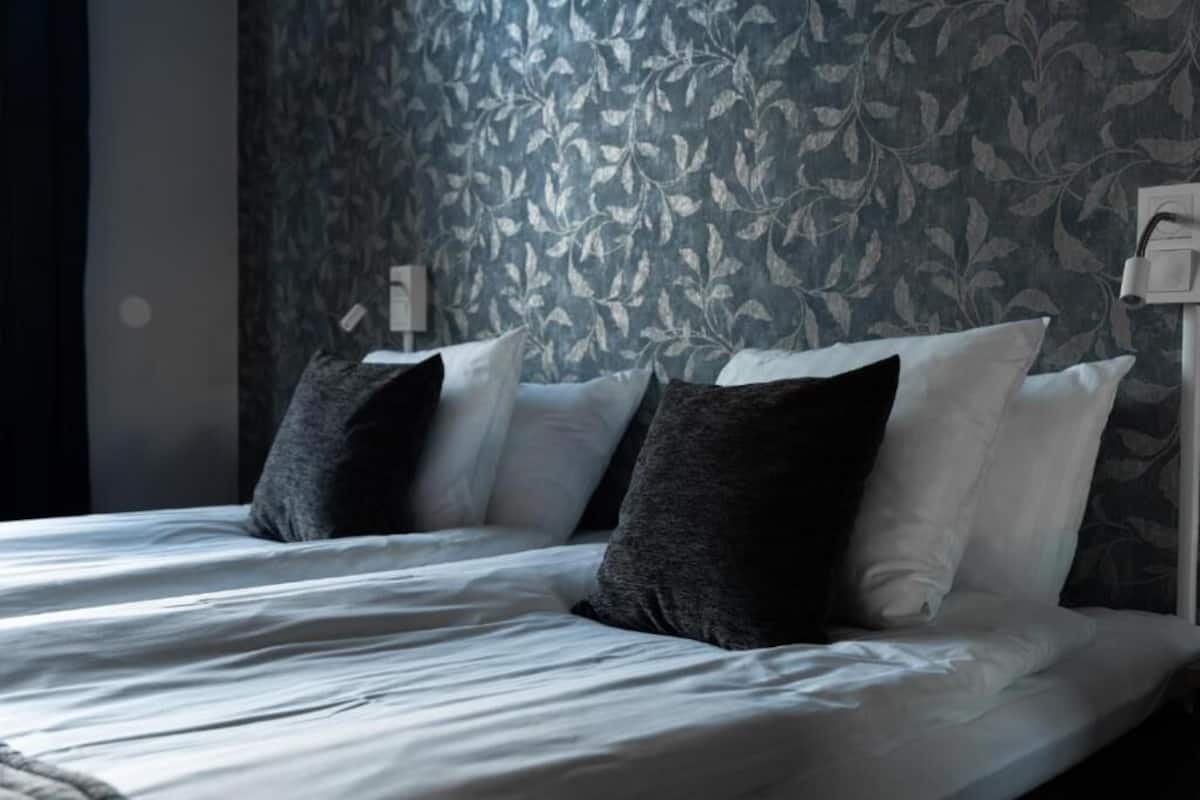 Cama bem arrumada com lençóis brancos e duas almofadas escuras, diante de uma parede com padrão de folhagens.