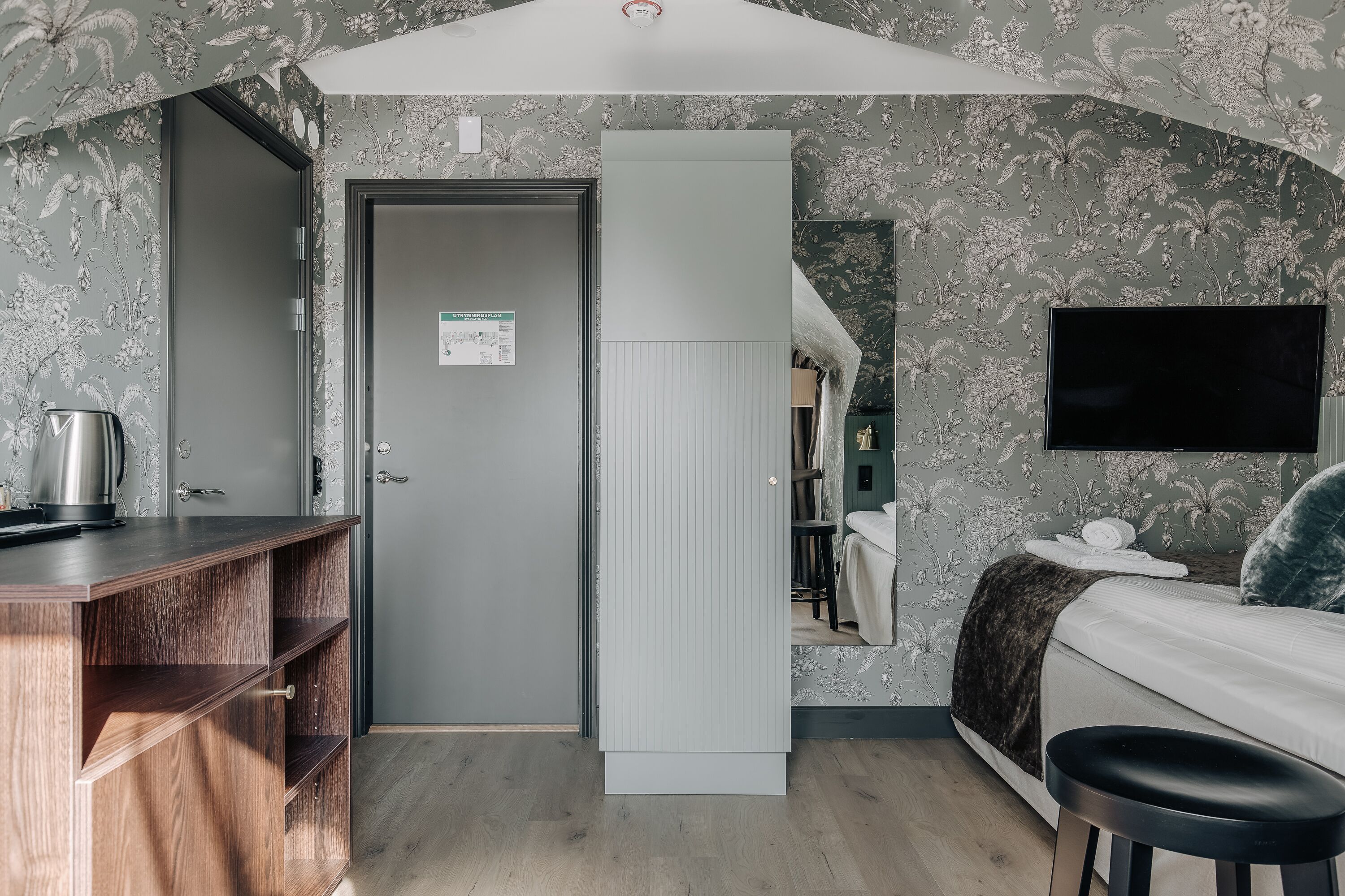 A room with a wooden desk, a bed with a dark blanket, a mounted TV, and a door with a sign.