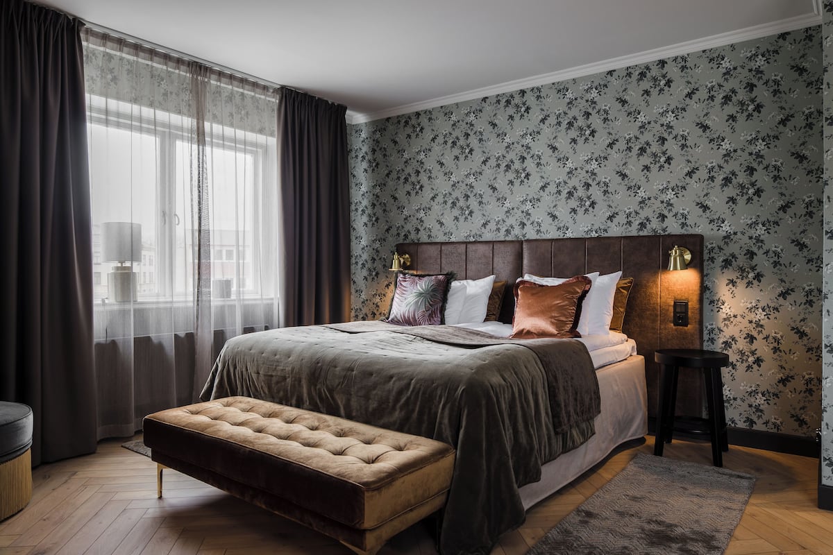 Quarto com cama grande, um banquinho, mesa de cabeceira e janela com cortinas.