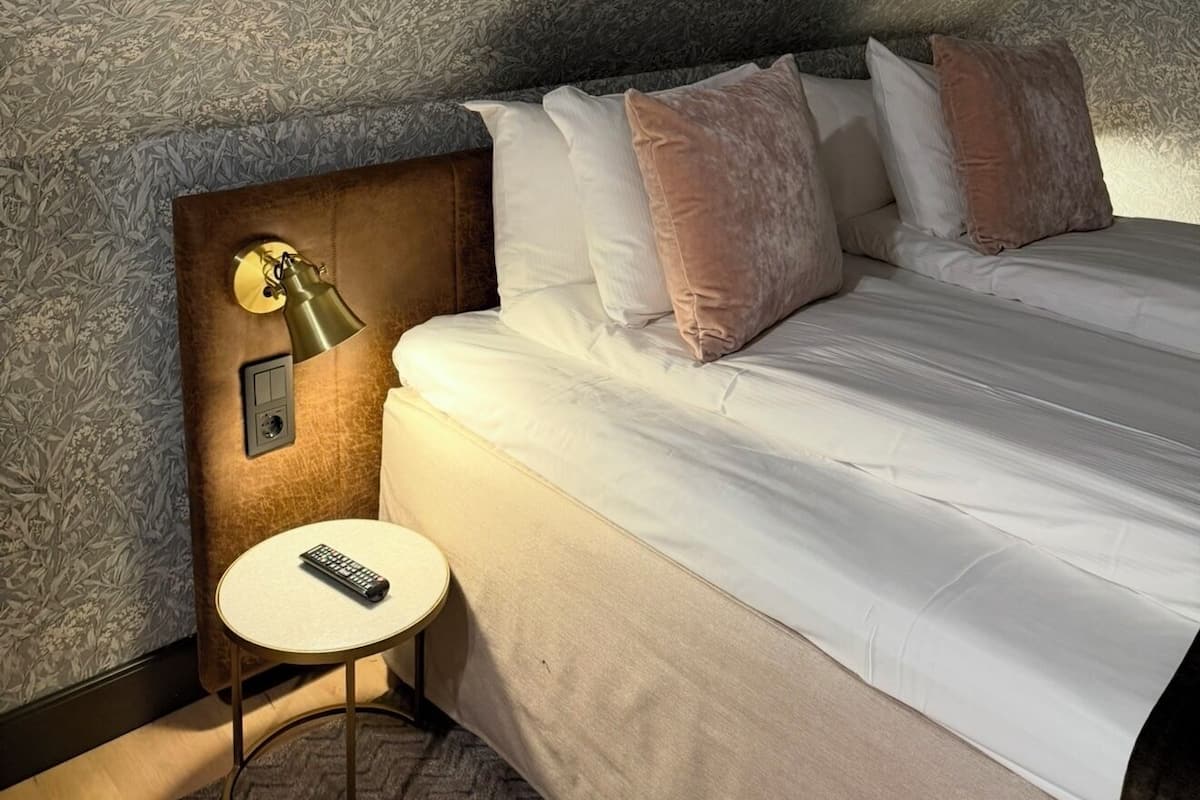 Quarto de hotel com cama, mesa de cabeceira, abajur e controle remoto sobre uma mesinha.