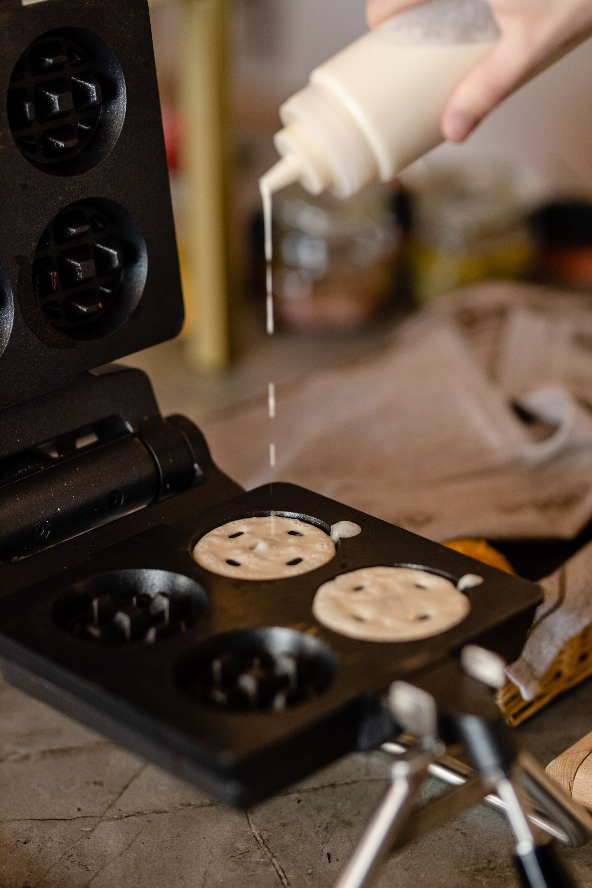 A waffle iron with four sections, a bottle being poured into it, and a wooden spoon nearby.