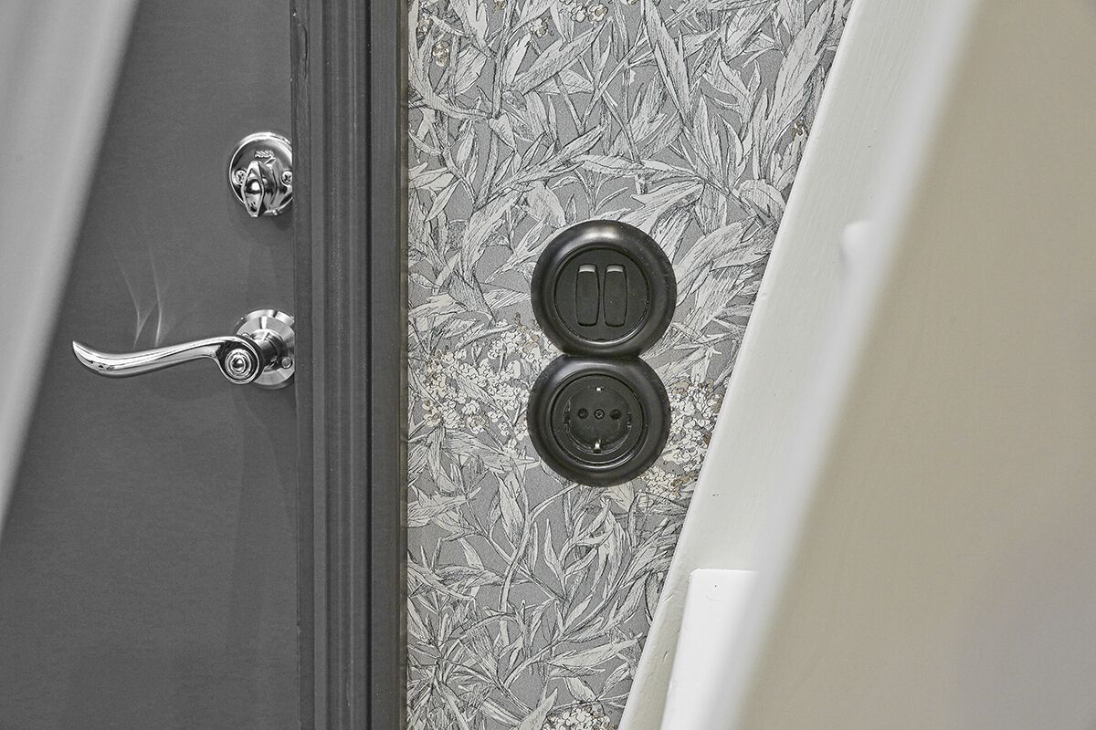 Uma porta com puxador prateado e olho de fechadura, e uma parede com papel de parede estampado com flores.