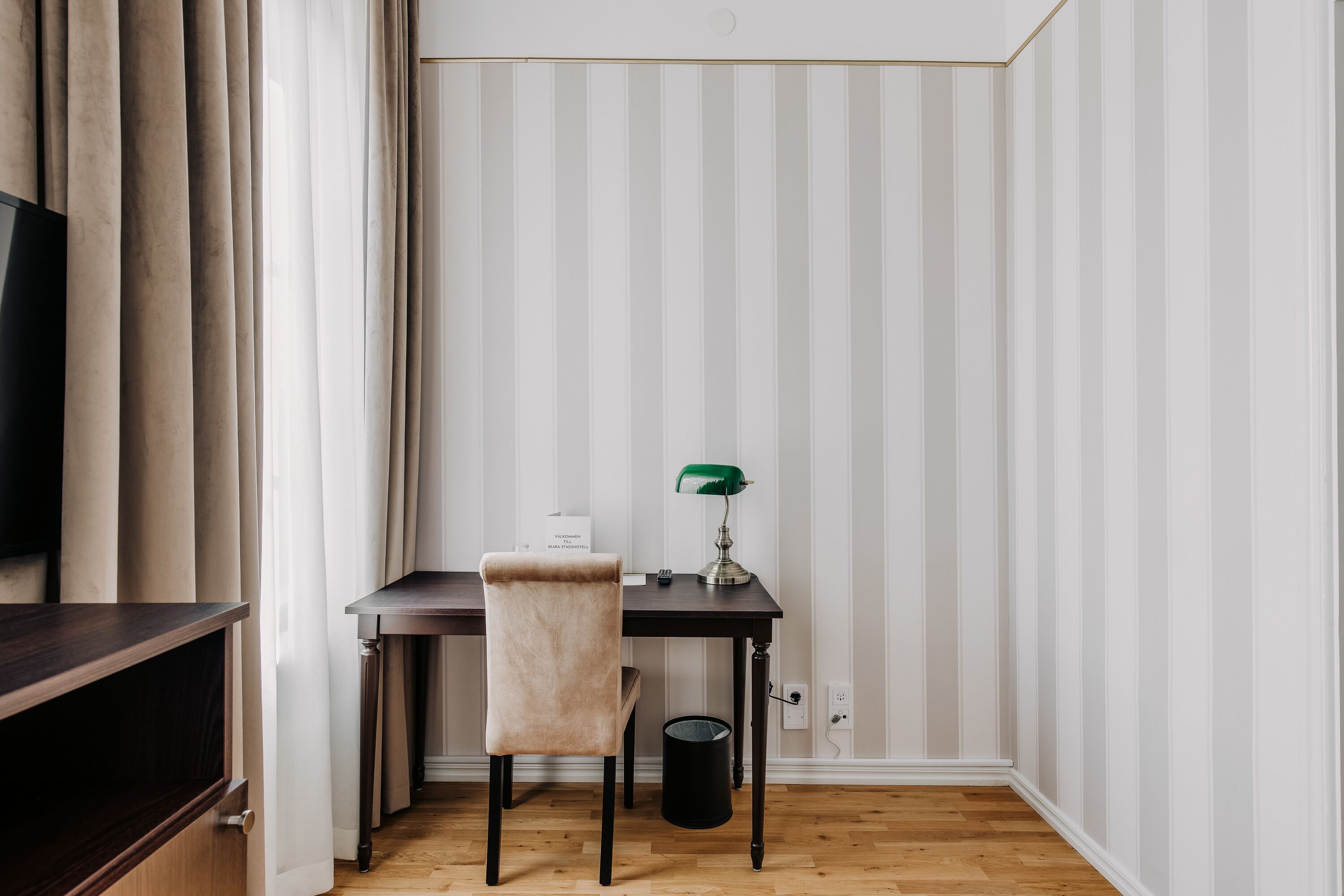 Een hoekkamer met een houten bureau, een stoel, een groene bureaulamp en een televisie links.