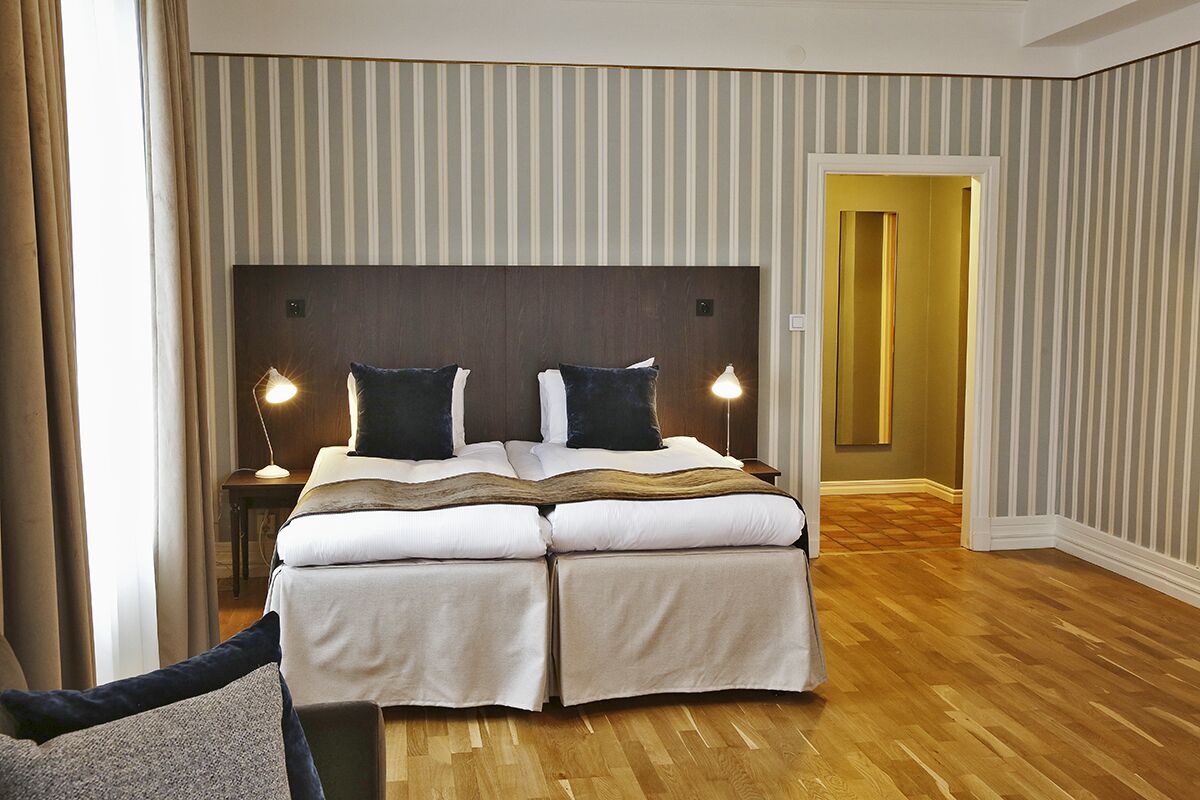 Habitación de hotel con una cama grande, dos lámparas de noche, un cabecero de madera y vista a un pasillo.
