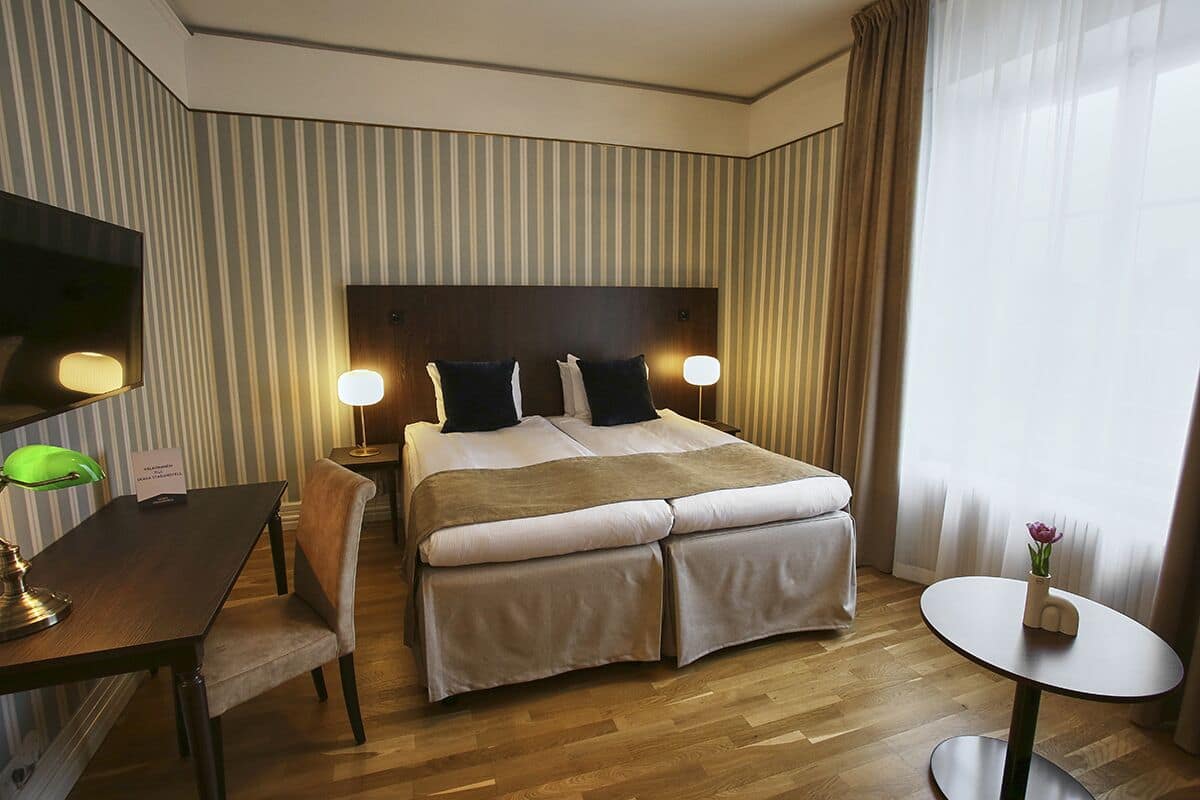 Quarto de hotel com cama, mesas de cabeceira, escrivaninha, cadeira, mesa redonda e televisão.