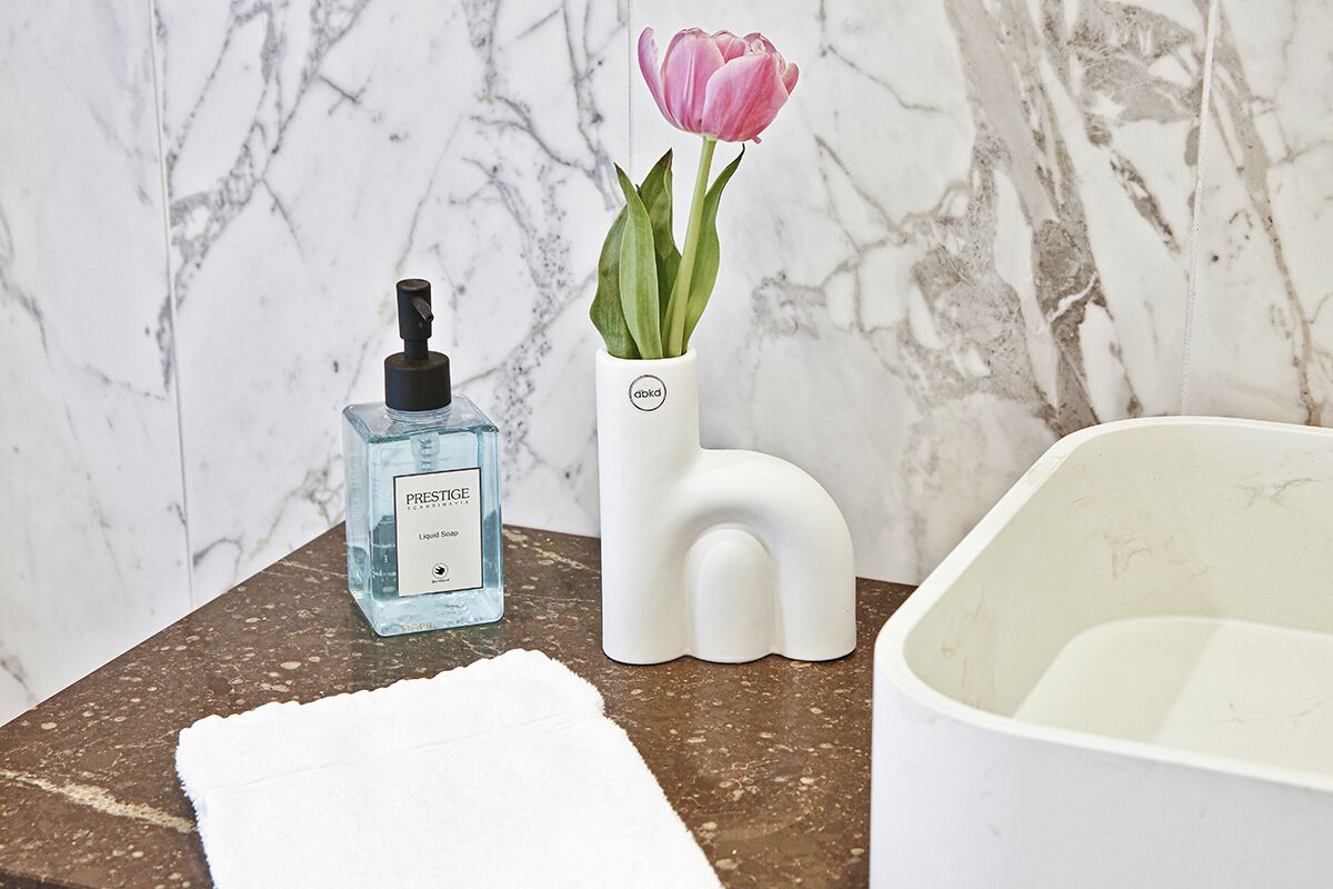 Un baño con pared de mármol, un jarrón blanco con una tulipán rosa, una botella de jabón líquido y una bañera blanca.