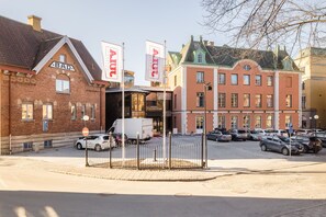 A street view with a row of buildings, including a red brick building with a 'BAD' sign, a white building with 'STADHOTEL' signage, and a yellow building. There are parked cars and a white van in front of the buildings. - Skara Stadshotell (Skara)