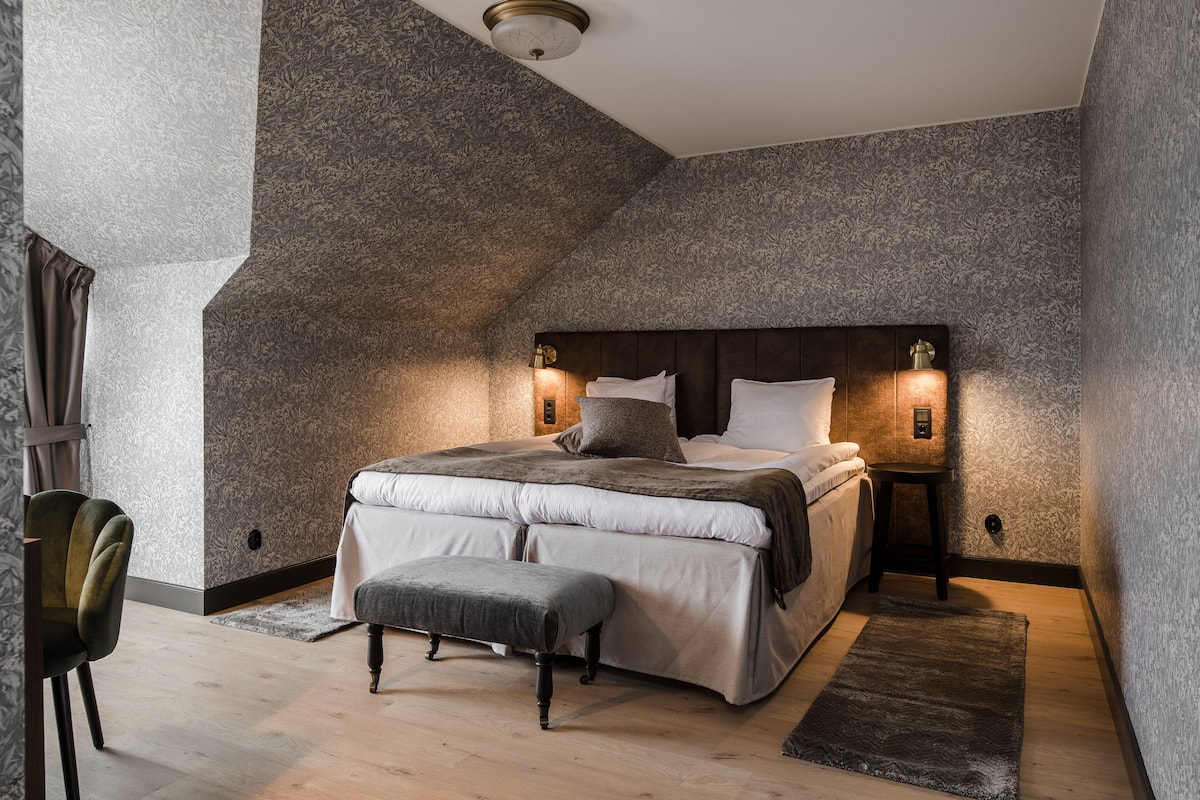Quarto com uma cama grande, duas mesas de cabeceira, um banquinho e uma cadeira.