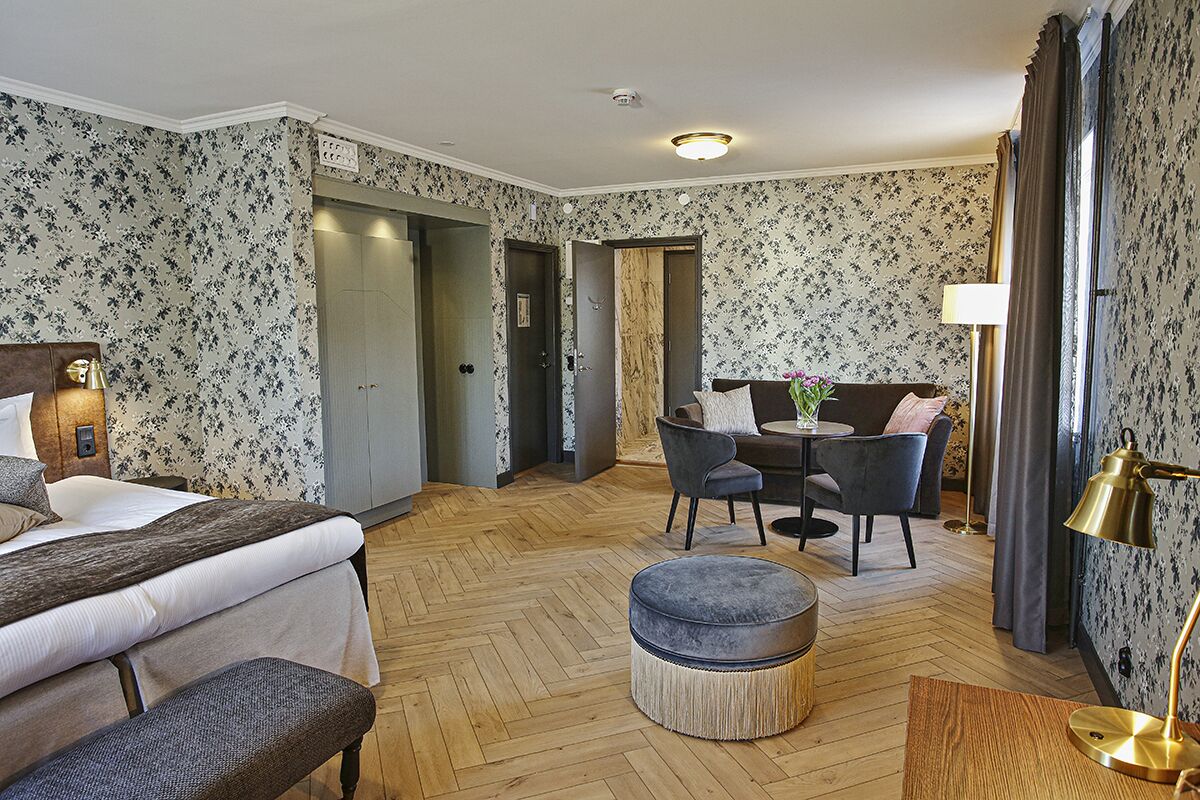A hotel room with a bed, a round ottoman, a small table with chairs, and a sofa. There is a lamp on the table and a lamp on a side table.