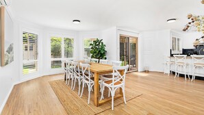 Uma ampla sala de jantar com mesa de madeira e cadeiras brancas, grandes janelas com persianas e vista para o exterior.