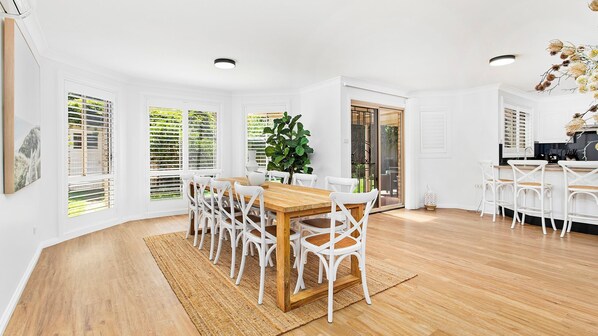 Un vaste espace dédié à la salle à manger, avec une table en bois et des chaises blanches, de grandes fenêtres équipées de stores et une vue sur l’extérieur.