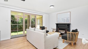 Uma sala de estar moderna com um sofá branco, uma cadeira de madeira, lareira e uma grande porta de vidro deslizante que dá para um jardim.