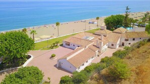 A coastal property with a beach view, a swimming pool, and a paved driveway leading to a white building with a tiled roof.