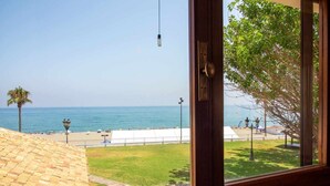 A view from a window overlooking a beach with a palm tree, a white fence, and a clear sky.