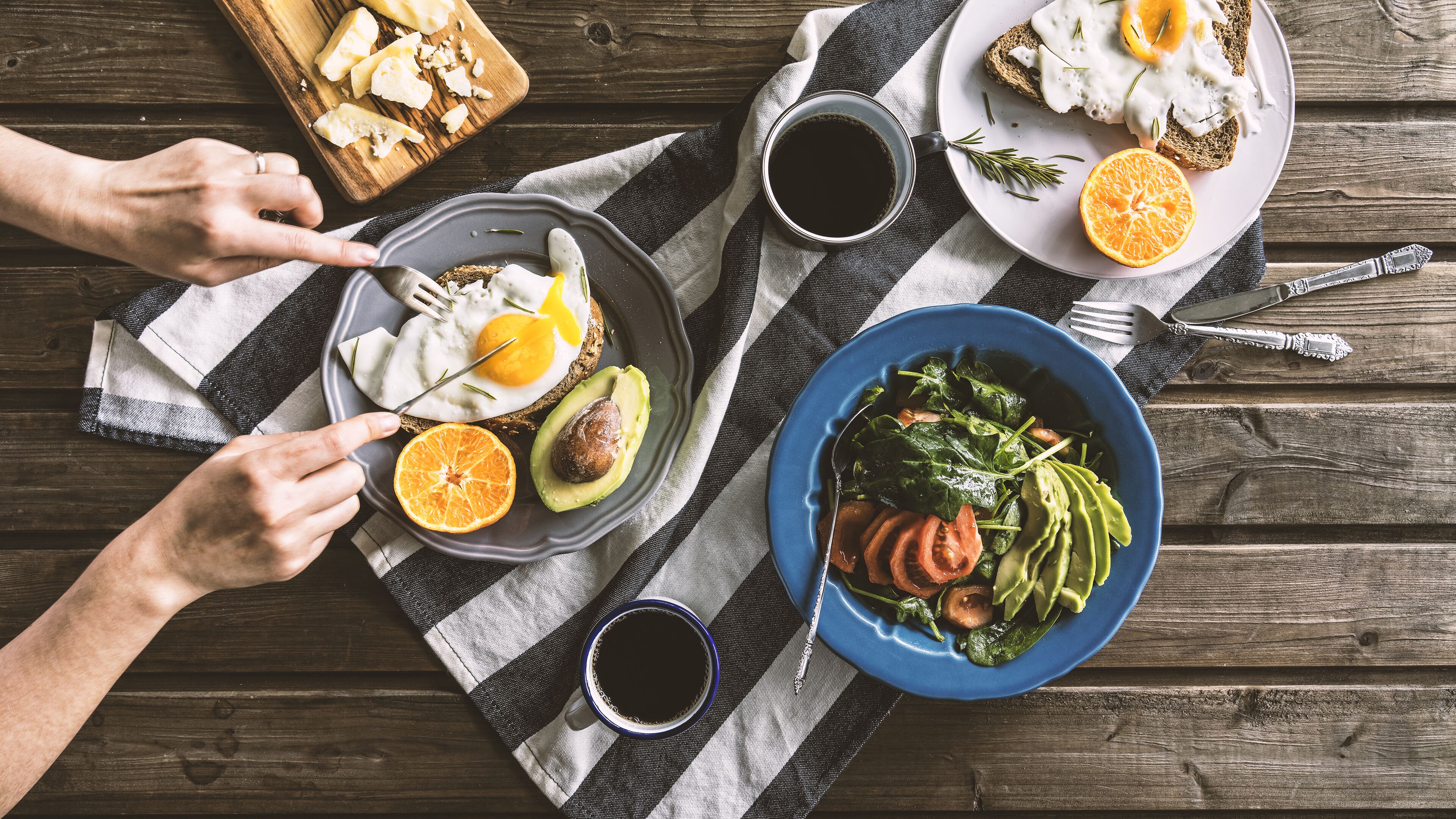 Desayuno con tostadas, huevos, aguacate, gajos de naranja y un bol de ensalada sobre una mesa de madera.