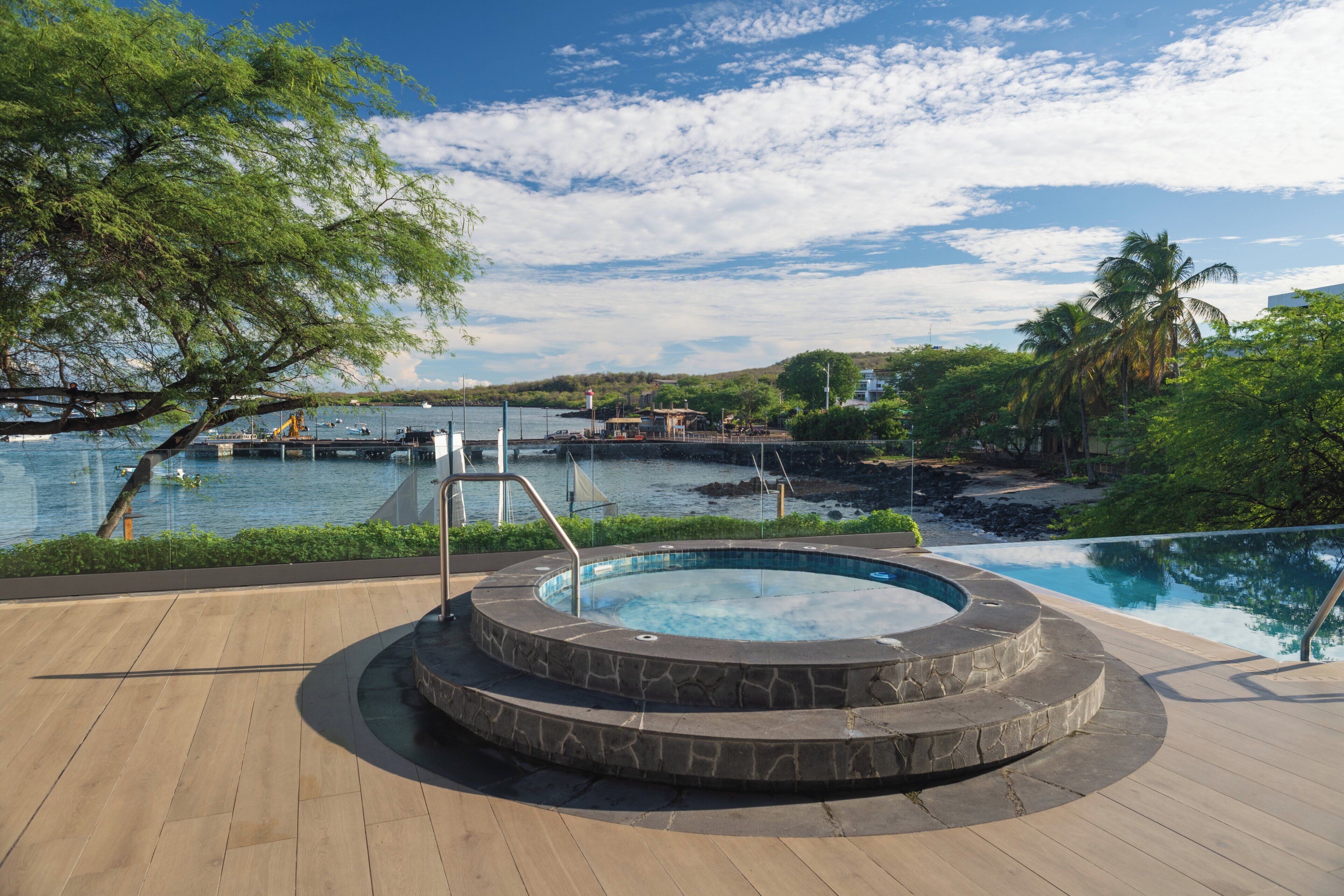 A hot tub surrounded by a wooden deck, overlooking a body of water with boats and a bridge in the background.