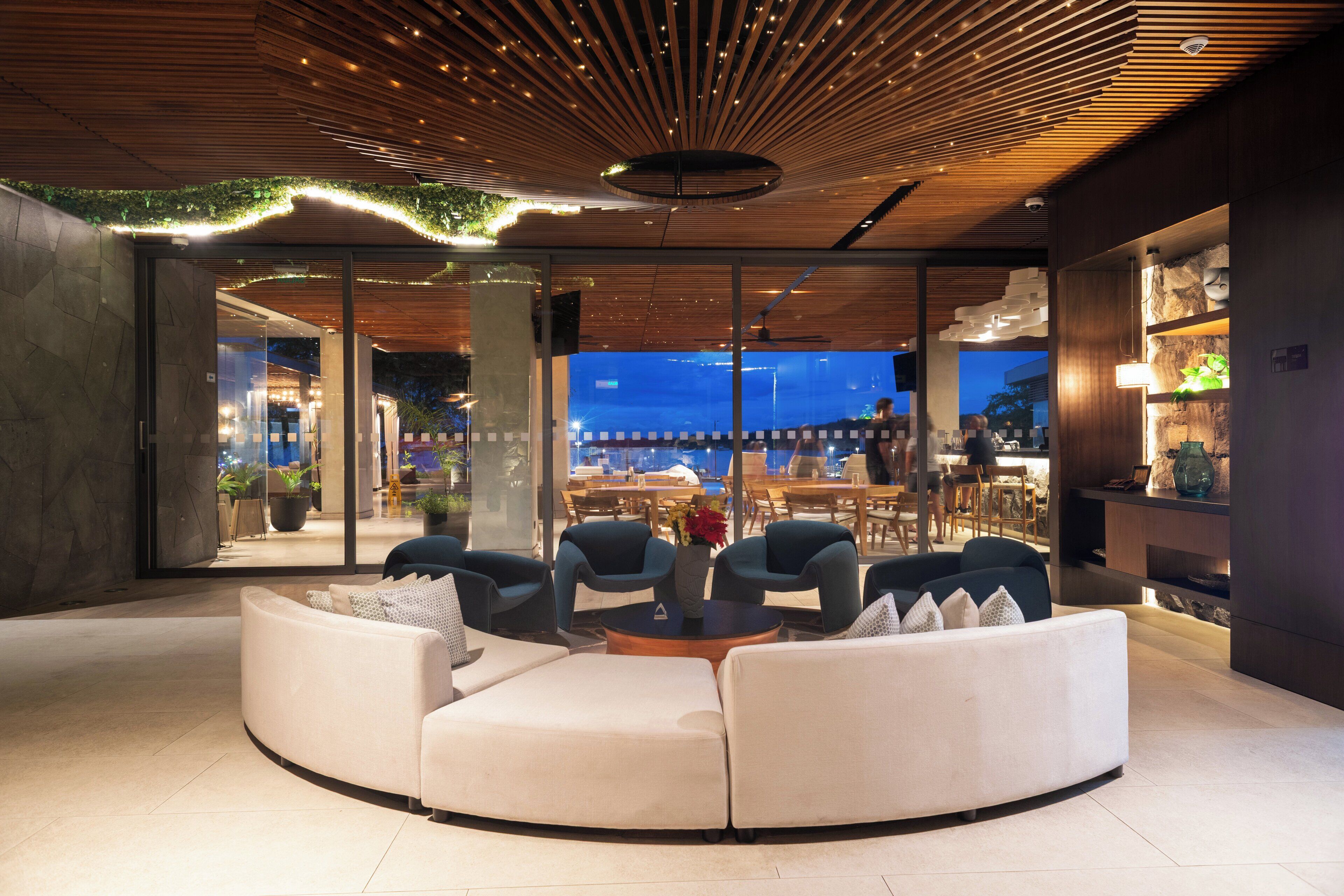 A modern lounge area with a curved sofa, a coffee table, and blue chairs. There is a large window with a view of the cityscape.
