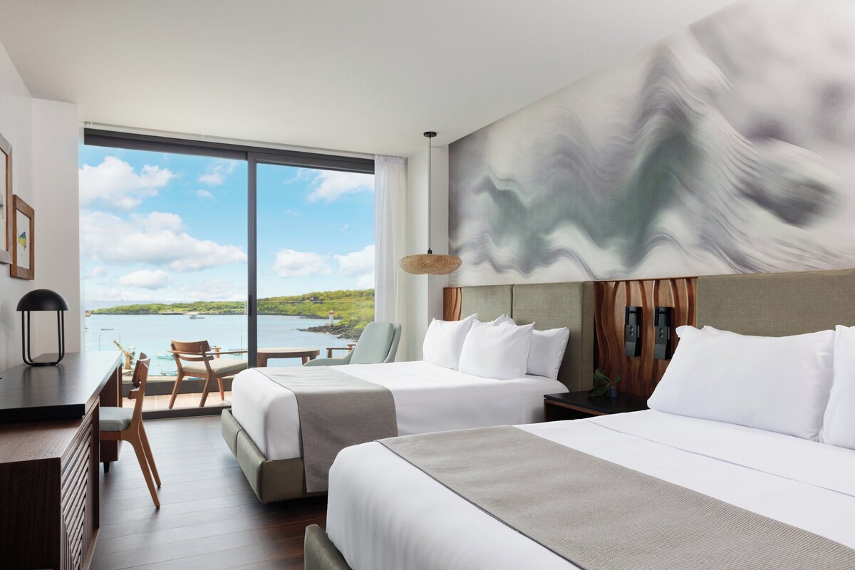 Ein Hotelzimmer mit zwei Betten, einem Schreibtisch und einem Stuhl. Es gibt ein großes Fenster mit Blick auf das Meer und die Landschaft.